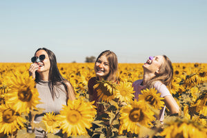 Three women smiling and laughing in a sunflower field wearing Nöz purple, orange, and blue zinc oxide sunscreen.